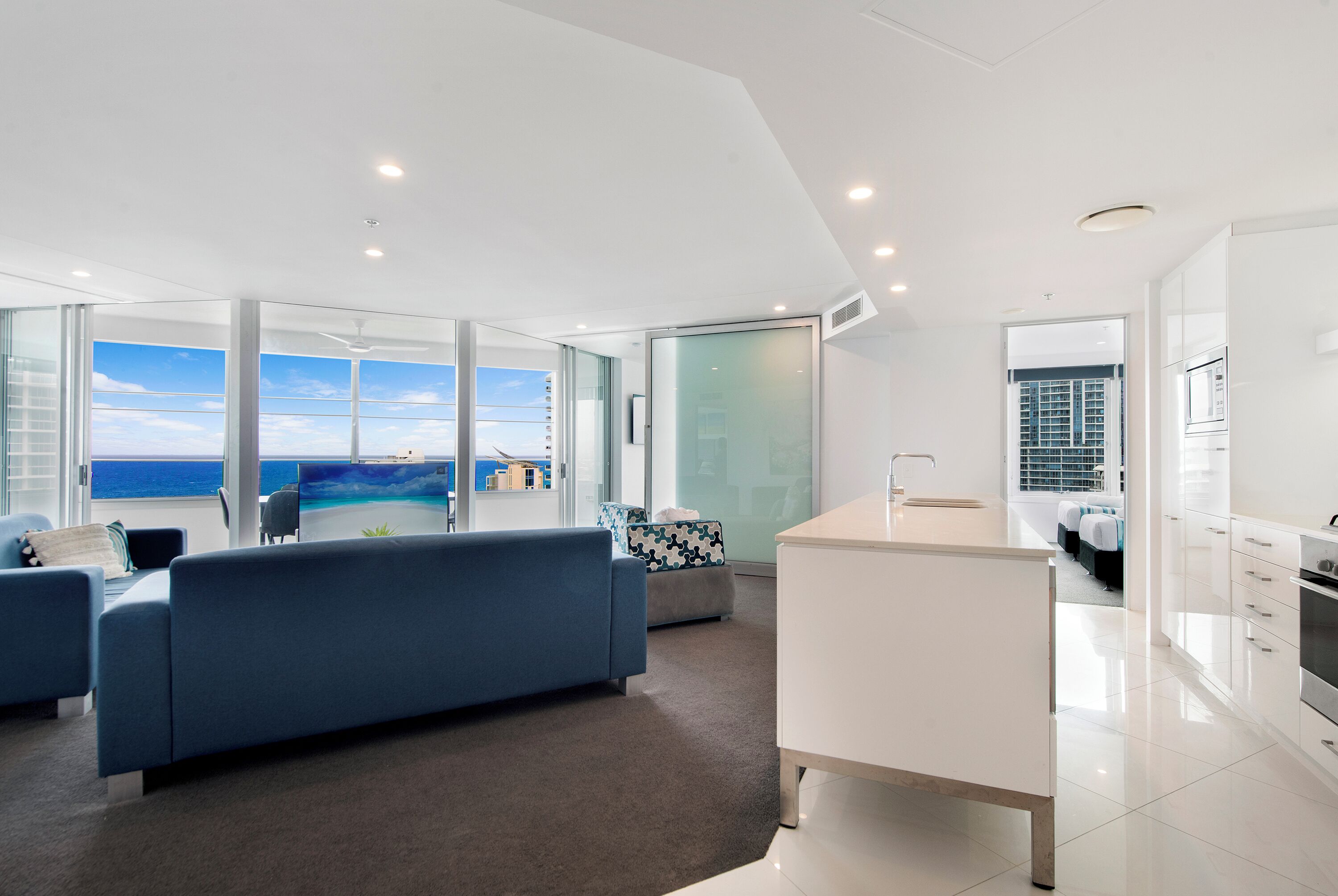 Una sala moderna con un ventanal grande con vista al océano, una cocina blanca con una isla central y un rincón de descanso acogedor.