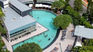 A rooftop pool with a curved design, surrounded by lounge chairs and umbrellas, and adjacent to a building with large windows and a sign. - Private Q Tower Apartments - WOWSTAY (Surfers Paradise)