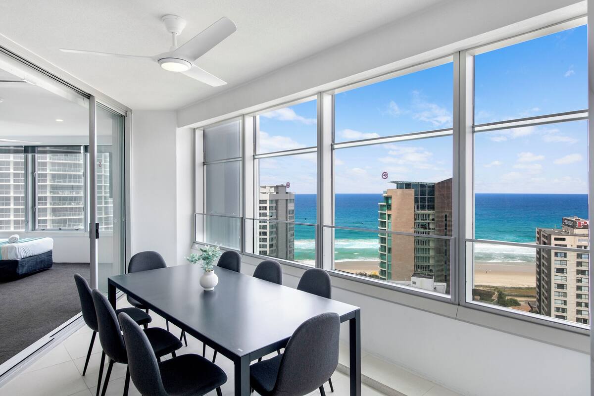 Une chambre d’hôtel moderne dotée d’un grand balcon, d’un coin repas avec une table et des chaises, et offrant une vue sur l’océan et les bâtiments.