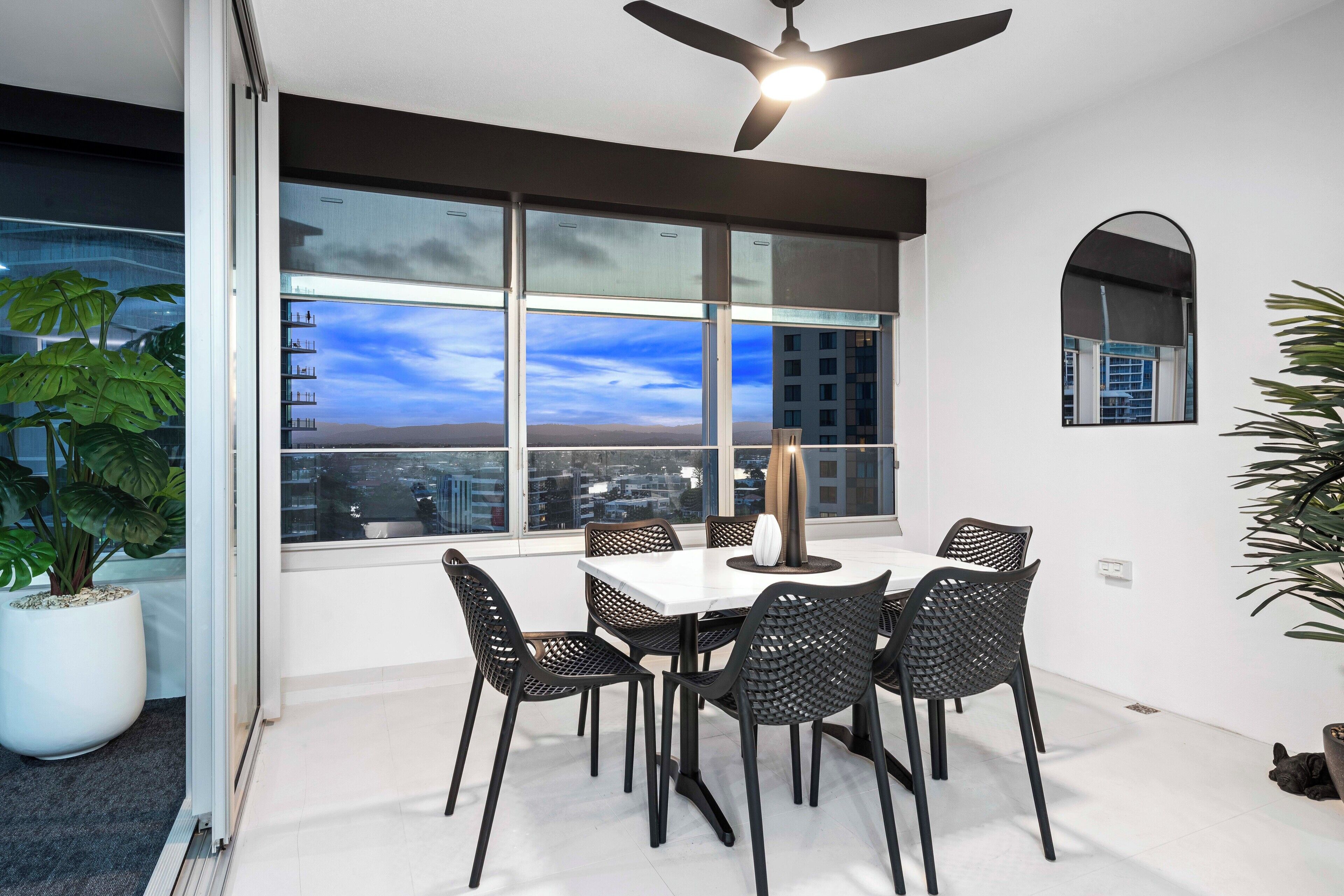 A modern dining area with a white table and black chairs, large windows with a city view, and a ceiling fan.