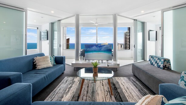 A modern living room with a large glass table, blue sofas, and a view of the ocean through large glass doors. - Private Q Tower Apartments - WOWSTAY (Surfers Paradise)