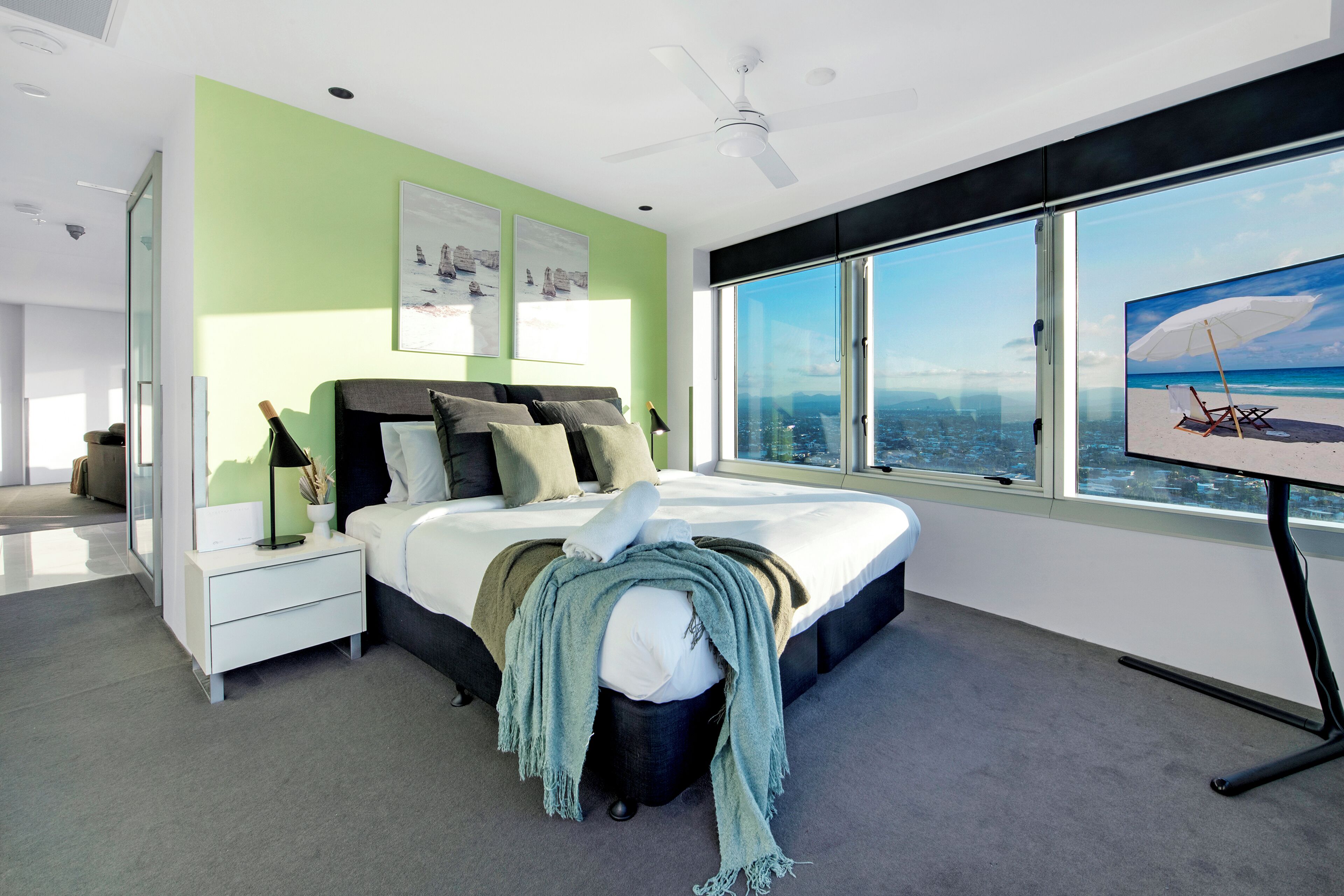 A modern bedroom with a large bed, a nightstand, a desk, and a view of the city through large windows.