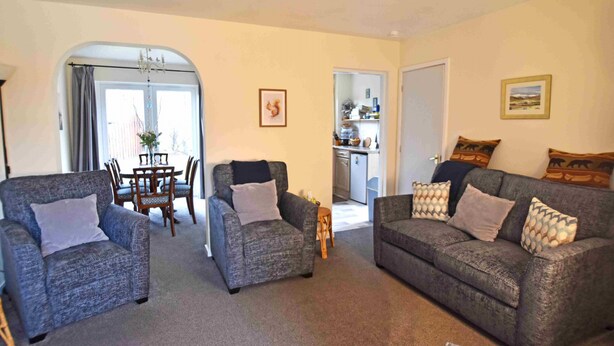 A living room with two grey armchairs, a grey sofa, and a dining table with chairs. There is a kitchen area visible through an open door. - Birch View -  Retreat Group (Aviemore)