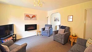 A living room with a flat-screen TV, two grey sofas, a blue armchair, a small wooden stool, and a chandelier. - Birch View -  Retreat Group (Aviemore)