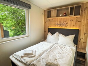 A neatly made bed with pillows and folded towels, a window with a view of greenery, and a wooden headboard with a shelf above.