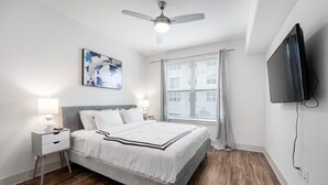 A bedroom with a bed, two bedside tables with lamps, a ceiling fan, a large window with blinds, a flat-screen TV mounted on the wall, and a painting on the wall.