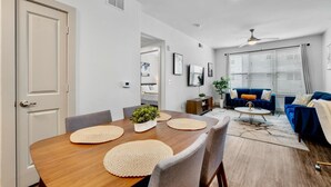 A modern living room with a dining area, featuring a wooden table, grey chairs, a blue sofa, and a ceiling fan.