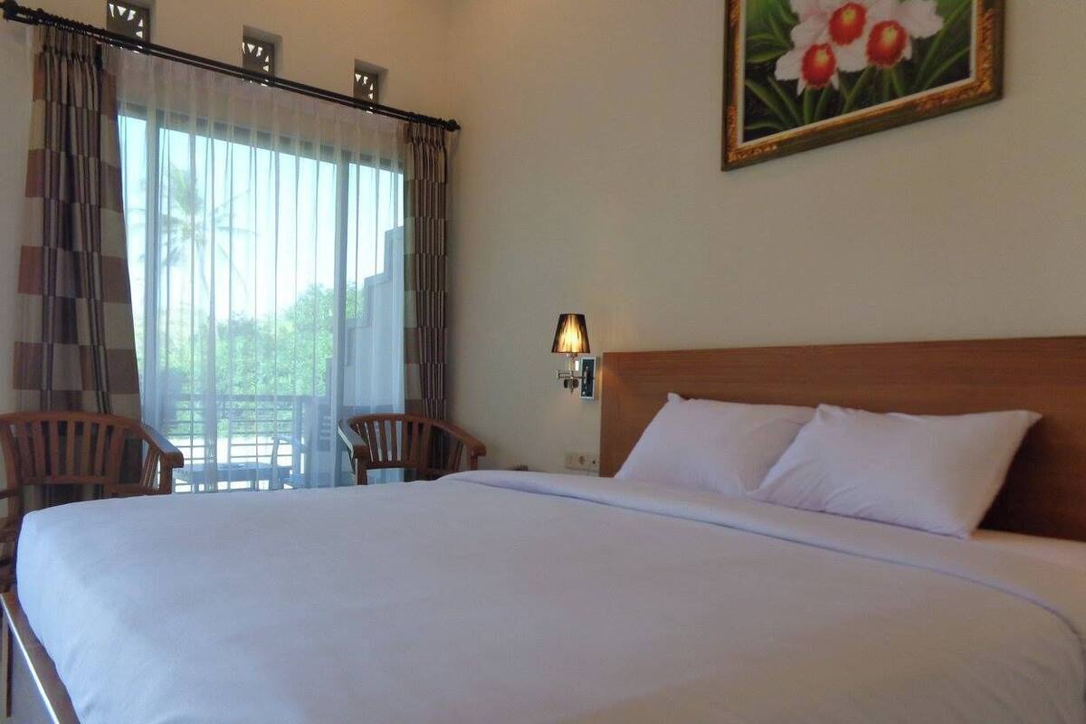 A hotel room with a large bed, a wooden headboard, a nightstand with a lamp, and a view through sheer curtains.