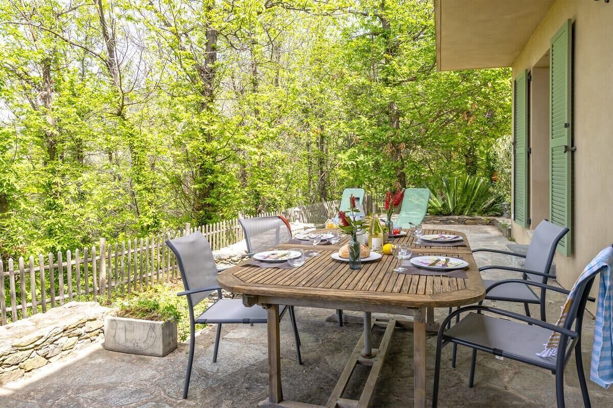Un espace extérieur aménagé pour les repas, avec une table et des chaises en bois, entouré d’un mur de pierre et d’une clôture en bois.