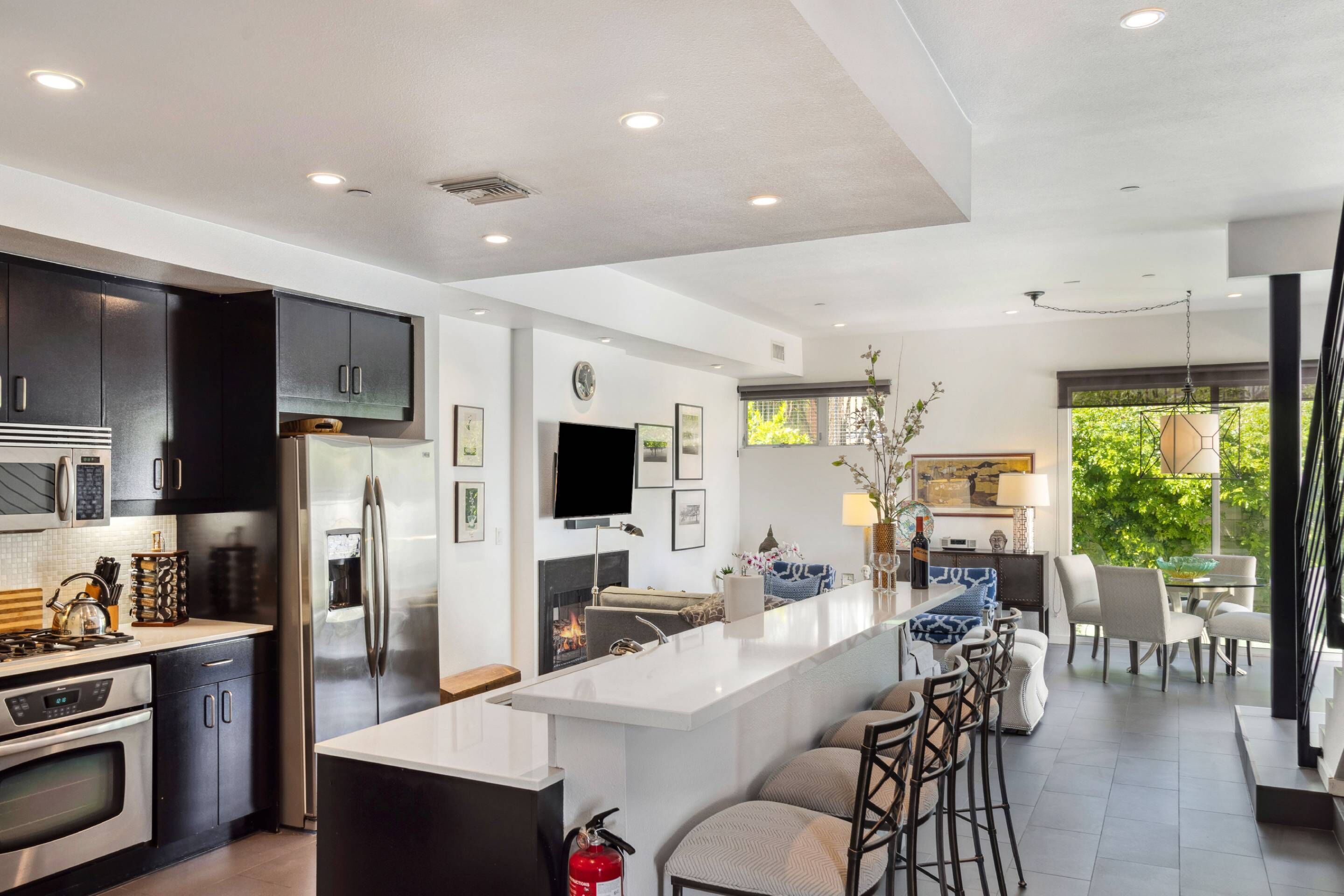 A modern kitchen with stainless steel appliances, a central island with bar stools, and a dining area with a view of greenery.
