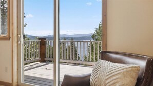 Une pièce avec un fauteuil en cuir marron, un coussin blanc et une porte-fenêtre coulissante donnant sur un balcon doté d’une terrasse en bois et offrant une vue sur les montagnes et un lac.