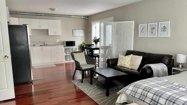 A modern living room with a black leather sofa, a dining table with chairs, a kitchen area with white cabinets, and a large window providing natural light. - Central Saanich cozy suite

 (Victoria)