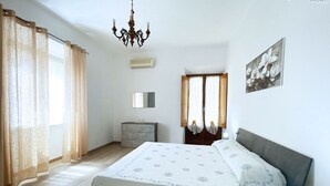 Een slaapkamer met een bed, een dressoir, een spiegel, een kroonluchter en een raam met gordijnen.