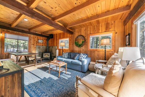 A cozy cabin interior with a wooden ceiling, a stone fireplace, a blue sofa, and a dining area.
