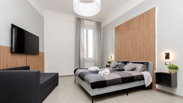 A modern bedroom with a bed, a sofa, a TV, a window with curtains, and a wall-mounted light. - VivimiHome - [Navigli-Duomo] Maison Fusetti (Milano)
