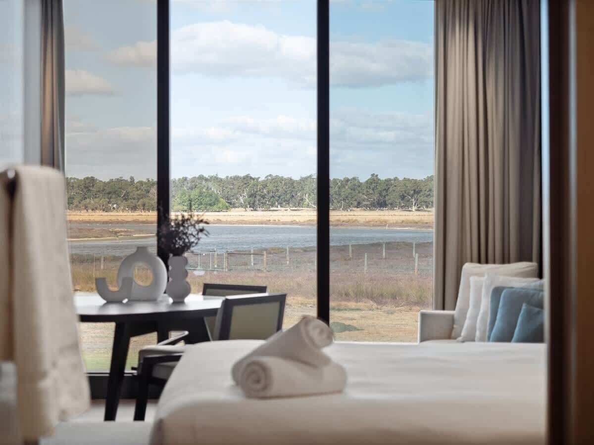 A hotel room with a large window, a bed, a table with a vase, and a view of a beach and water.