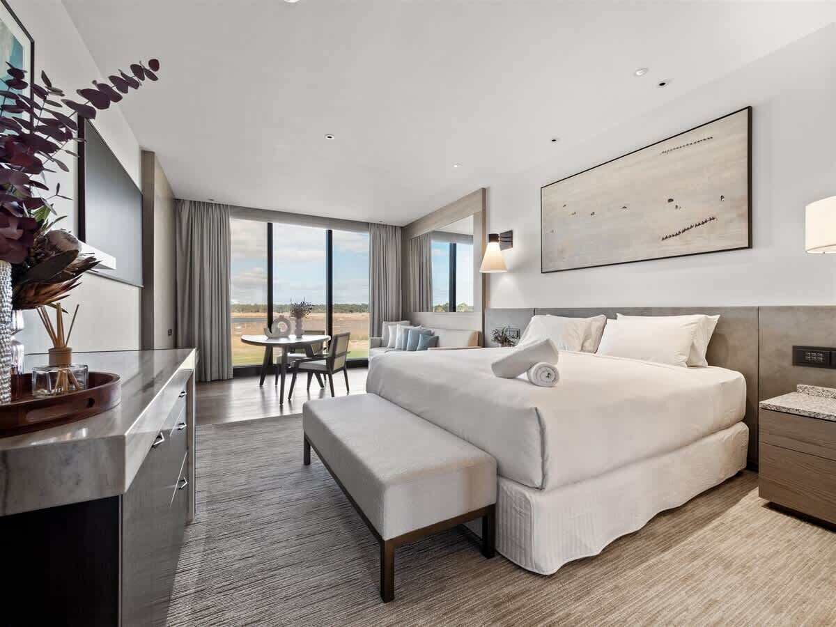 A modern hotel room with a large bed, a desk, a bench, and a view of the outdoors.