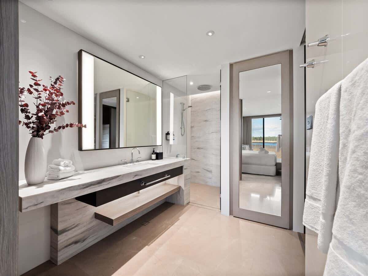 A modern bathroom with a large mirror, a vanity with a marble countertop, a shower area, and a view of the outdoors.