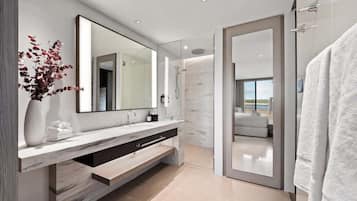 A modern bathroom with a large mirror, a vanity with a marble countertop, a shower area, and a view of the outdoors.
