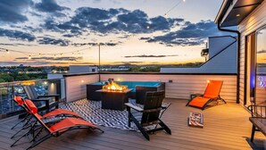 En tagterrasse med bålsted, liggestole og lyskæder mod en solnedgang.