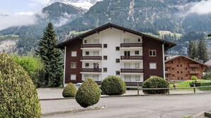 A multi-story building with balconies, surrounded by lush greenery and mountains in the background.