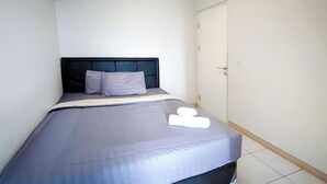 A neatly made bed with a grey bedspread and pillows, a white towel on the bed, and a closed door to the right. - Summarecon Springlake Basella  (Bekasi)
