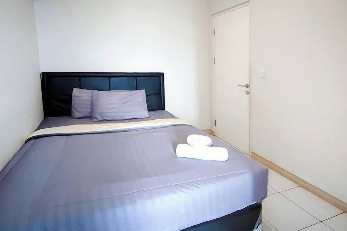 A neatly made bed with a grey bedspread and pillows, a white towel on the bed, and a closed door to the right.