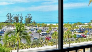 Vista da janela com vista para um estacionamento, palmeiras e uma praia ao fundo.