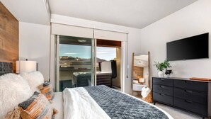 Ein modernes Hotelzimmer mit einem großen Bett, einem Flachbildfernseher, einer dunklen Holzkommode und einer Glastür, die auf einen Balkon mit Blick auf die Stadt führt.