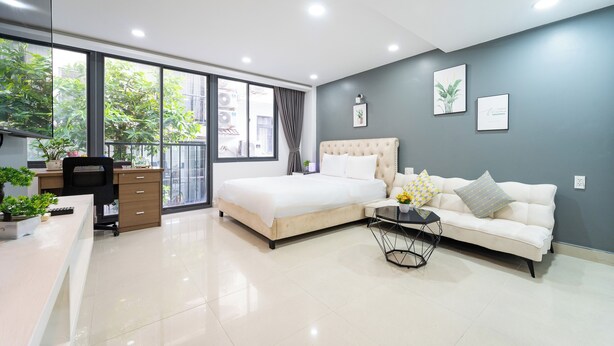 A modern bedroom with a bed, a sofa, a desk, and a large window. - Golden Suites 3 Apartments (Ho Chi Minh City)