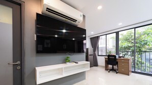 A modern office space with a mounted air conditioner, a mounted flat-screen TV, a white desk with a black chair, and a large window offering a view of greenery outside. - Golden Suites 3 Apartments (Ho Chi Minh City)