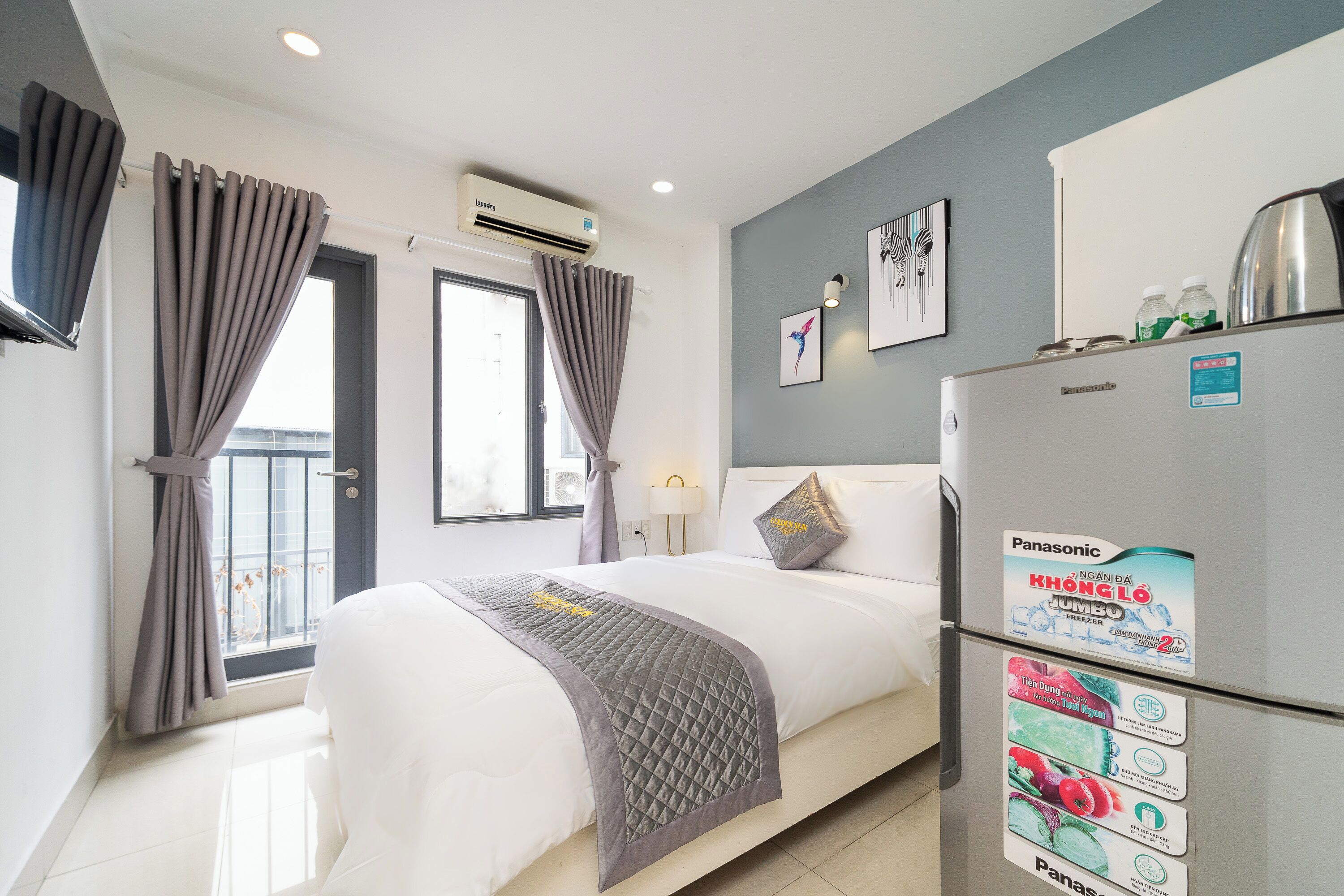 Een hotelkamer met een bed, gordijnen, een raam, een airconditioner, een koelkast en wanddecoratie.