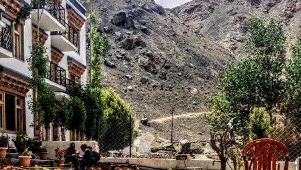 Un patio con un sendero bordeado de flores amarillas, una silla roja y un edificio con balcones y plantas en macetas.