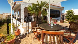Eine Terrasse mit rundem Tisch, Stühlen und einem großen Sonnenschirm. Übertöpfe mit Pflanzen und Blick auf ein Gebäude mit weißer Fassade und Terrakotta-Dachziegeln.