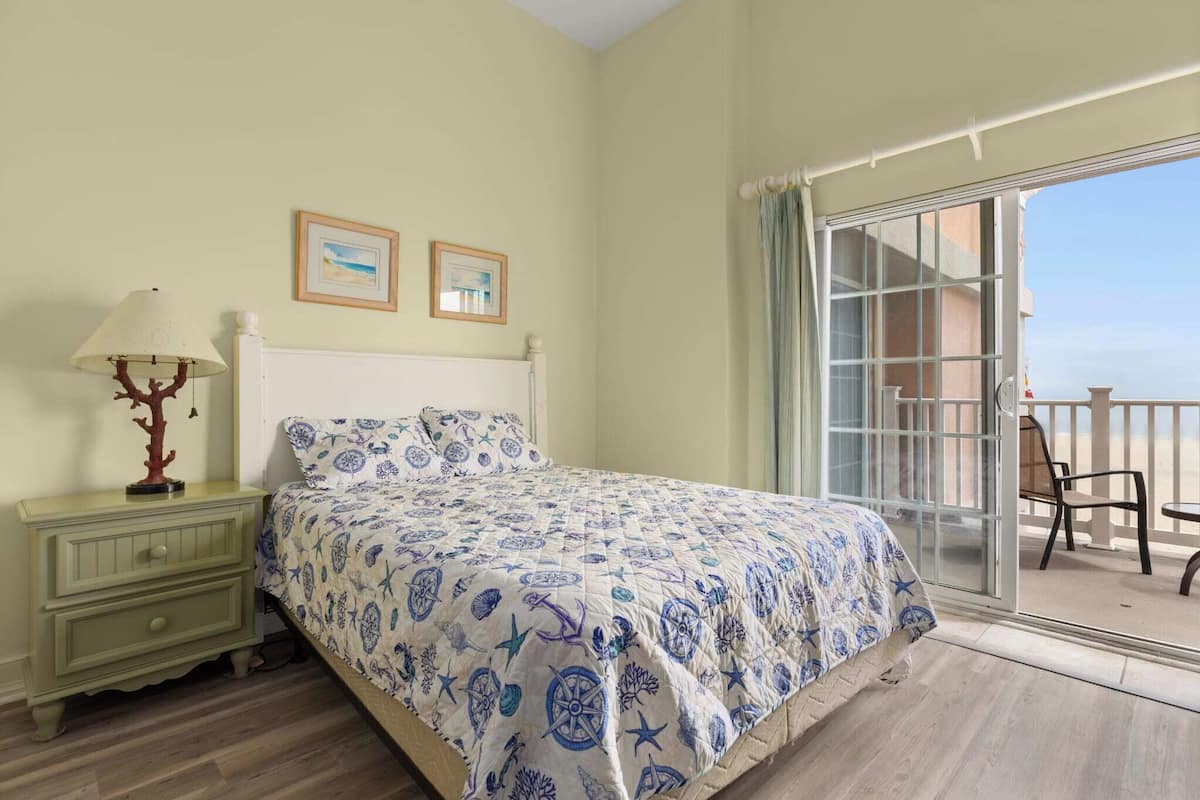 A bedroom with a bed, a nightstand, a lamp, and a view of the balcony.