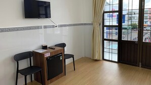 Una habitació amb televisió penjada a la paret, un escriptori petit amb un ordinador i un cartell, dues cadires i una finestra amb vista al carrer.