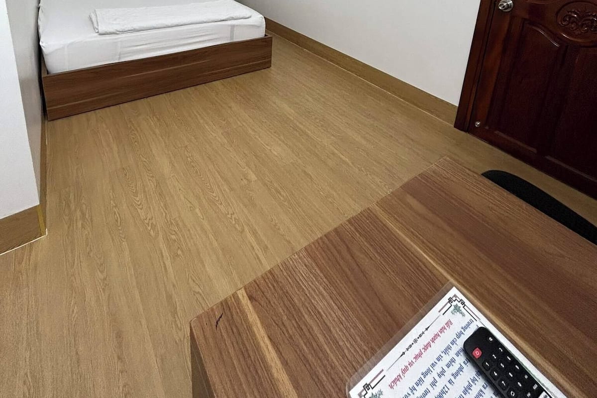 A bedroom with a wooden bed, white walls, a wooden door, and a small table with a remote control.