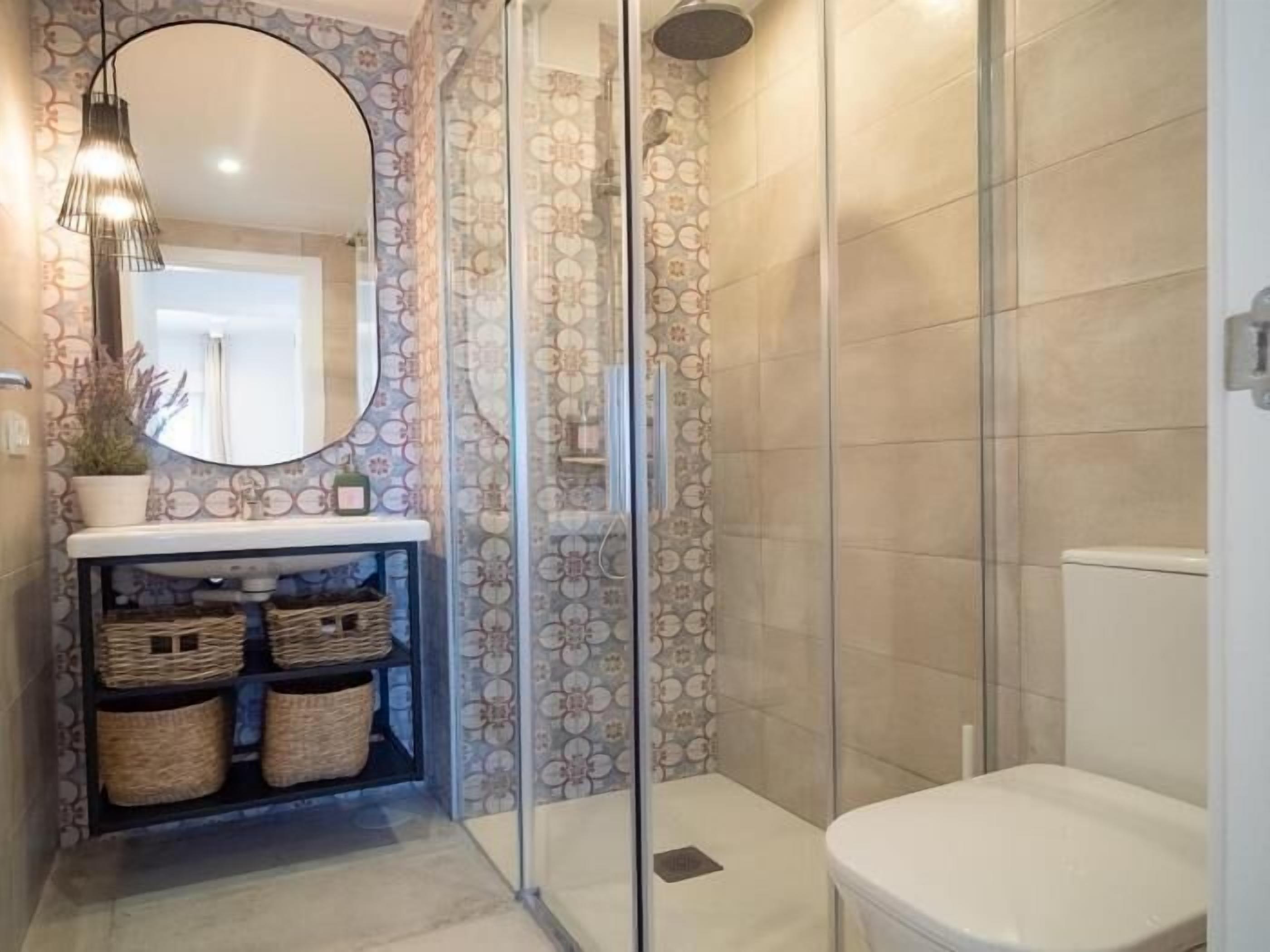 A bathroom with a glass shower enclosure, a round mirror, and a sink with wicker baskets.
