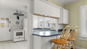 Una cocina con gabinetes blancos, horno doble, microondas y pileta. Hay dos taburetes de madera y una ventana con persianas.
