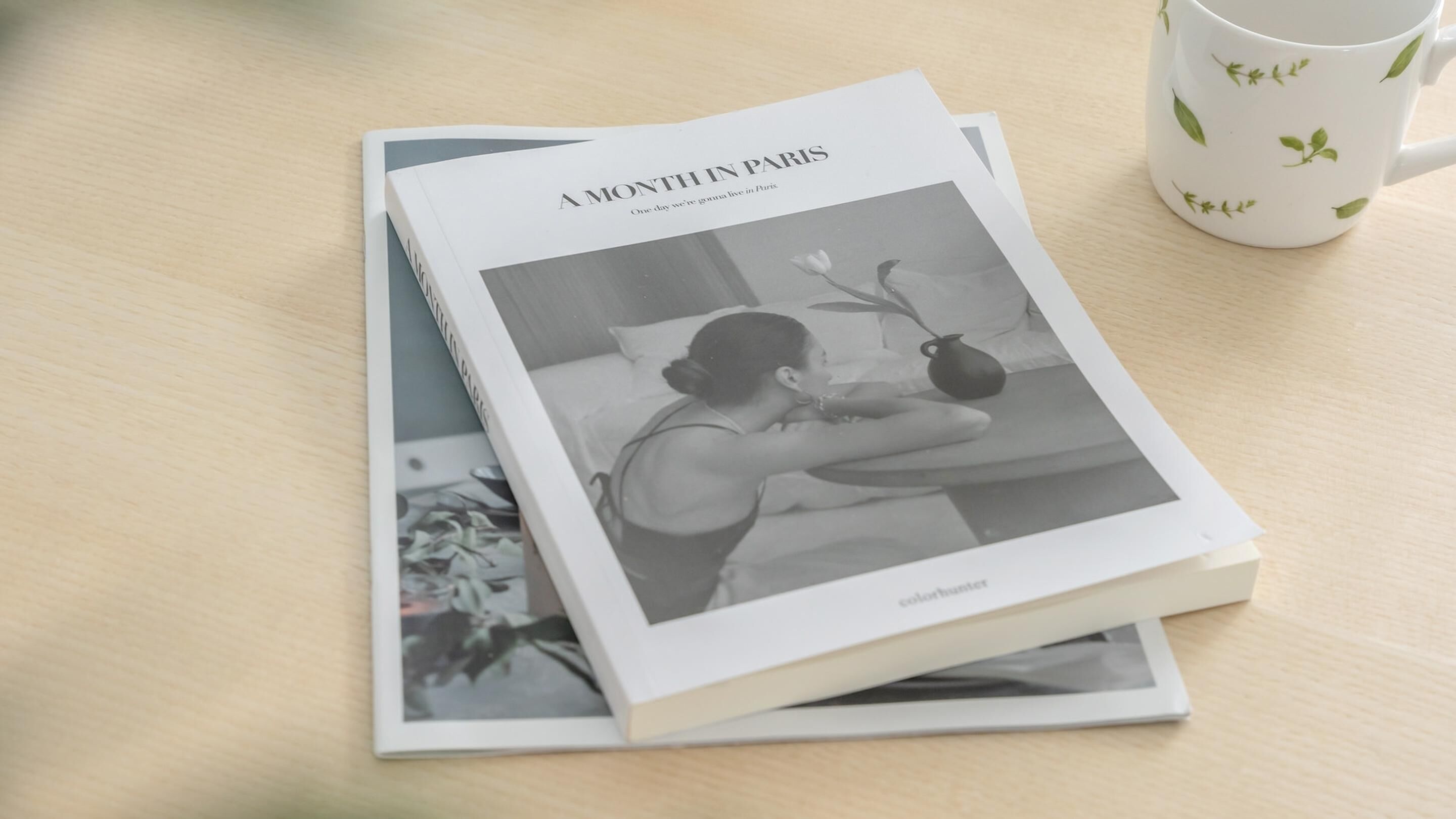 Un libro titulado 'A Month in Paris' con una fotografía en blanco y negro en la portada, sobre una superficie de madera al lado de una taza blanca con diseños de hojas.