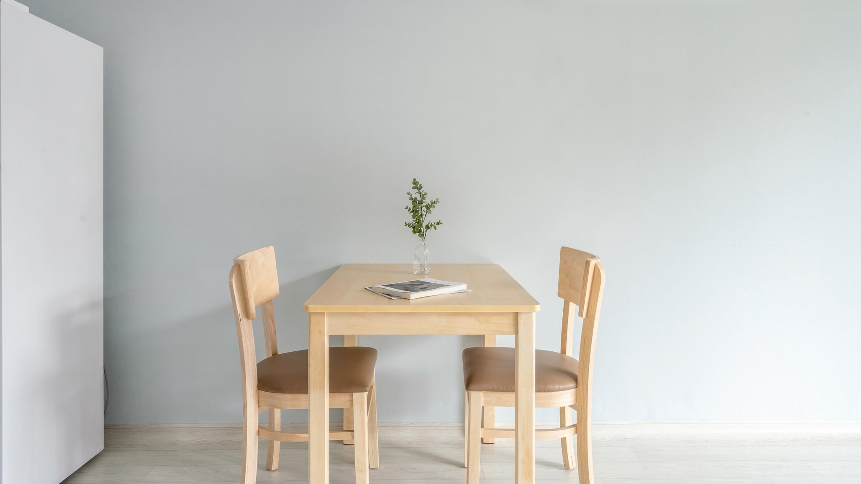 A minimalist dining area with a wooden table and two chairs, a small vase with a plant, and a book on the table.