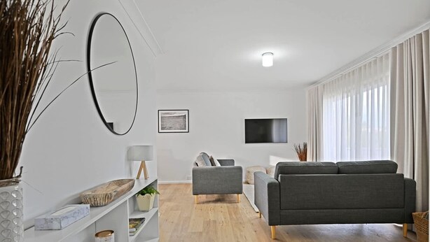 Een moderne woonkamer met een grijze bank, een salontafel en een televisie aan de muur. Er is een boekenplank met decoratieve items en een grote spiegel.