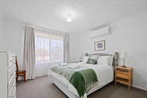 A hotel room with a bed, a nightstand, a dresser, a window with curtains, and an air conditioning unit.