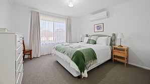 Een slaapkamer met een bed, een kaptafel, een stoel, een nachtkastje met een lamp, en een airconditioner aan de muur.
