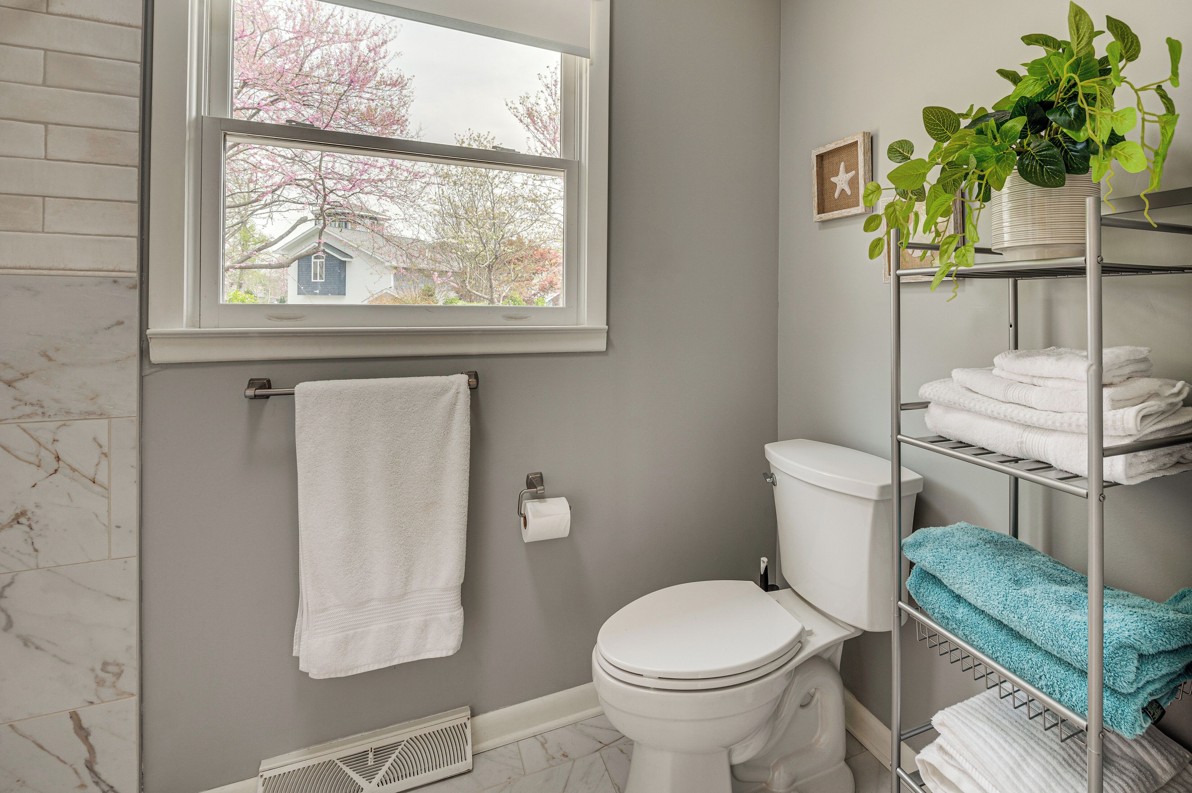 Ein Badezimmer mit Toilette, einem Handtuchhalter mit weißem Handtuch, einer Ablage mit gefalteten Handtüchern und einem Fenster mit Blick auf Bäume und ein Haus.