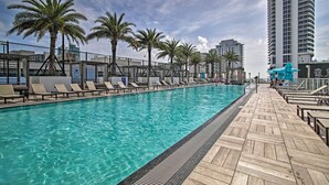 A swimming pool with lounge chairs and palm trees, surrounded by a wooden deck and a tall building in the background. - Walk to Hollywood Beach: Upscale 16th-Floor Condo (Hollywood)