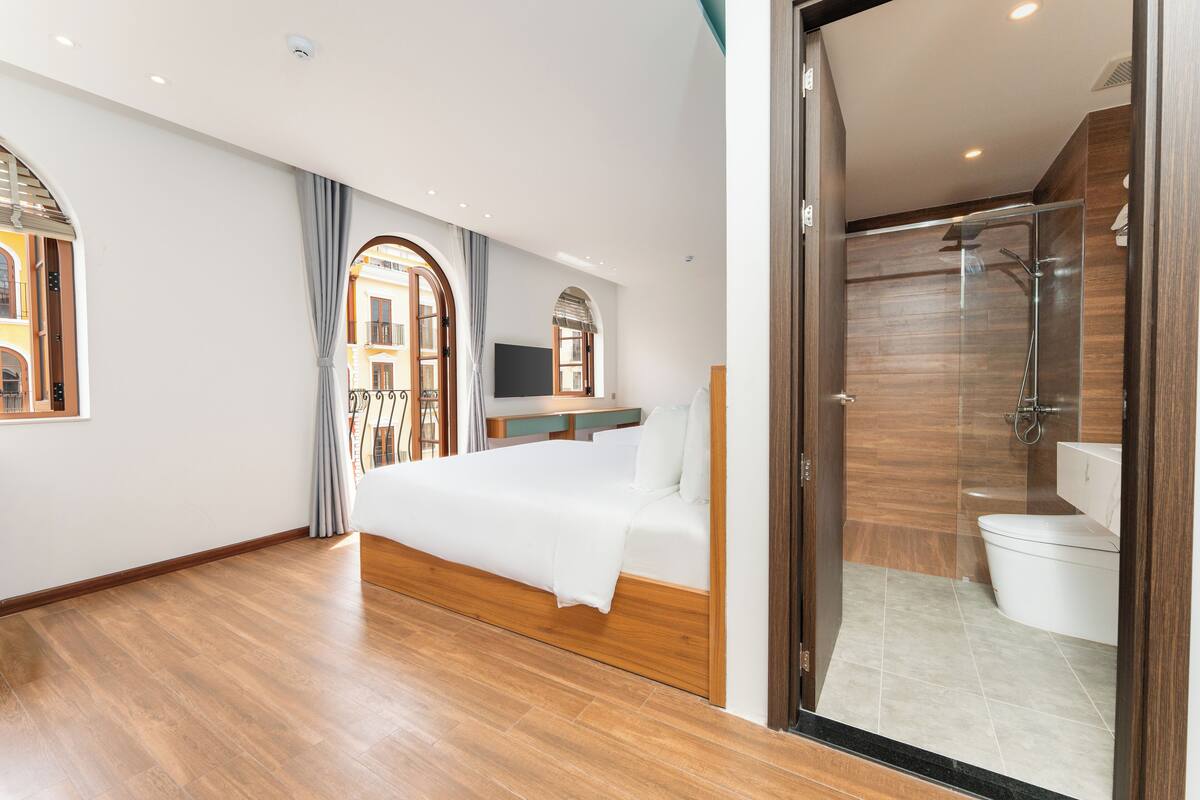 A modern hotel room with a wooden floor, a bed, a TV, and a glass-enclosed shower area.