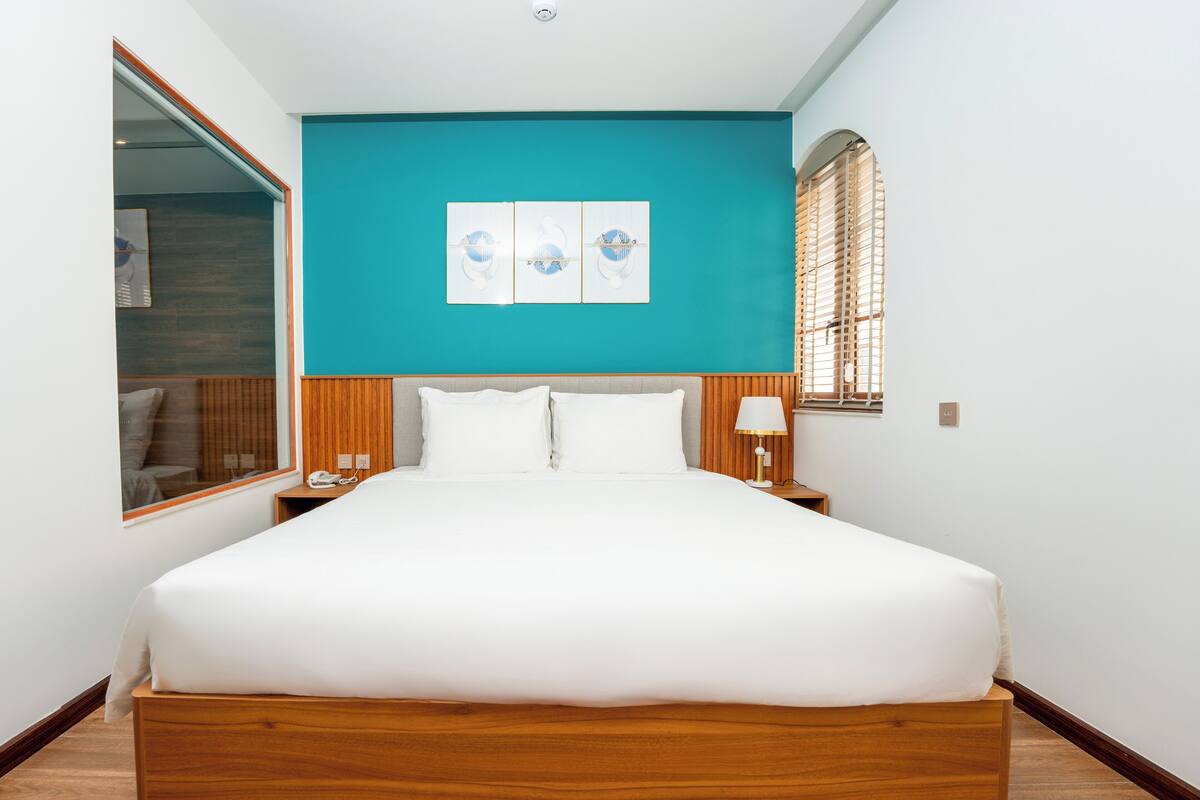 A hotel room with a large bed, a wooden headboard, a teal accent wall, a window with blinds, and a nightstand with a lamp.