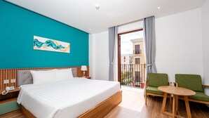 Ein Hotelzimmer mit einem großen Bett, einem Holznachttisch, einem Telefon, einem grünen Sessel, einem kleinen runden Tisch und einem Balkon mit Blick auf Gebäude.