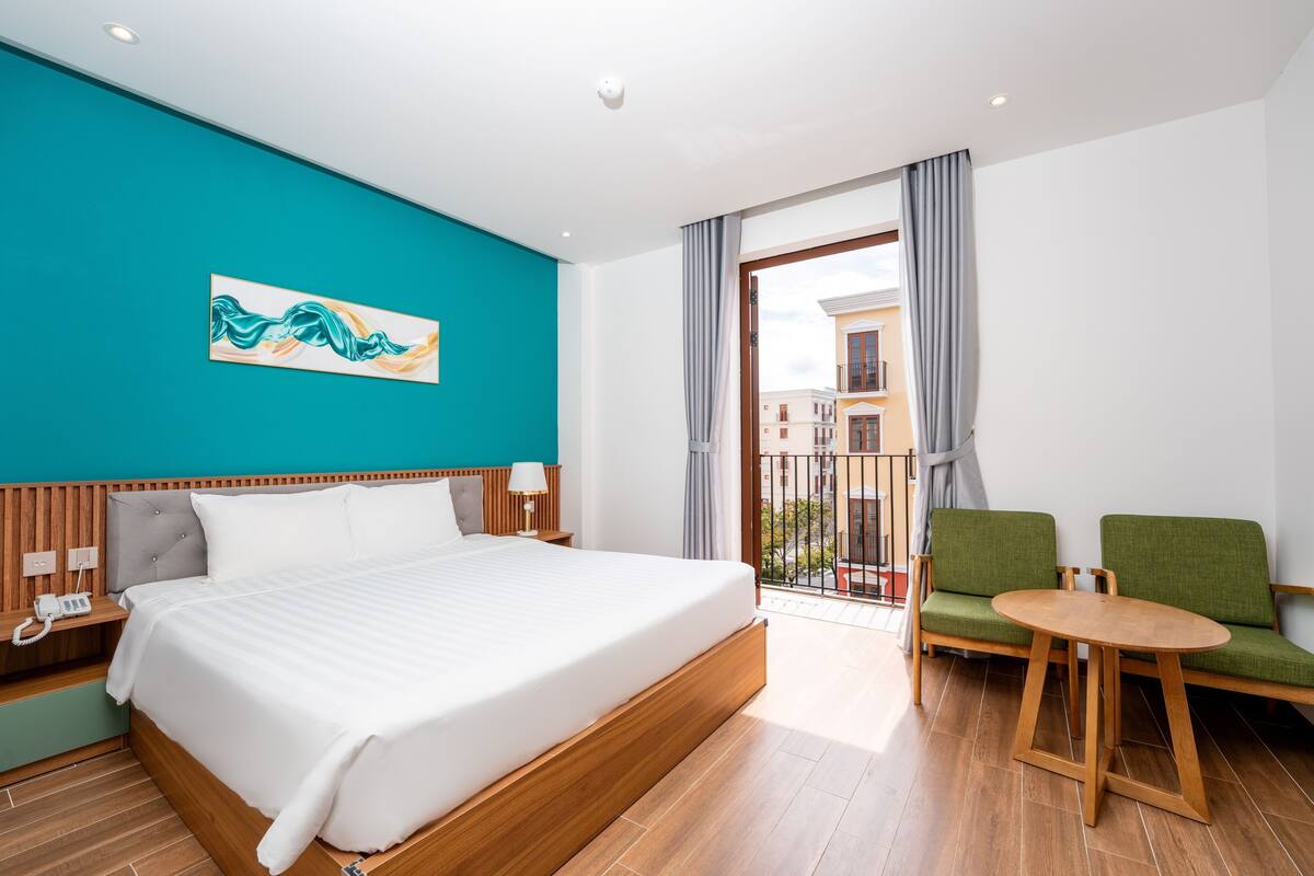 A hotel room with a large bed, a wooden nightstand, a telephone, a green chair, a small round table, and a balcony with a view of buildings.